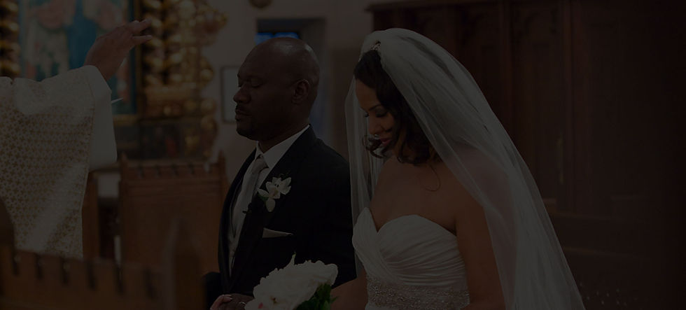 A bride and groom in their wedding clothes being blessed by a priest